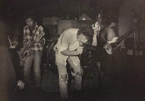 A black and white photo of a band performing live, with a singer in a torn shirt and two guitarists on stage.