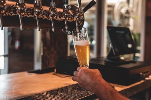 A hand holds a glass of beer at a bar, with beer taps and a register in the background.