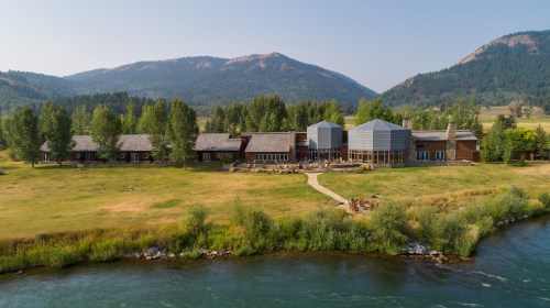 A scenic view of a large lodge by a river, surrounded by trees and mountains under a clear sky.