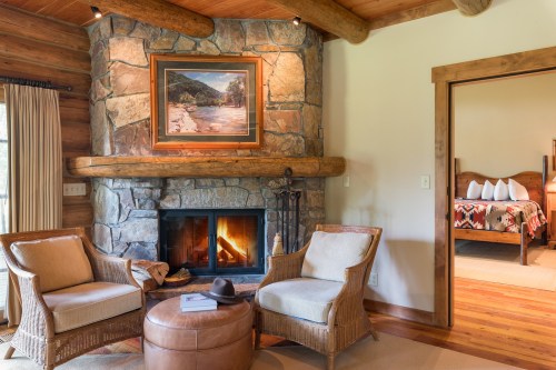 Cozy cabin interior with a stone fireplace, two chairs, and a view into a bedroom with a rustic bed.