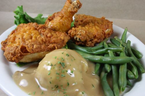 A plate of fried chicken with green beans and mashed potatoes topped with gravy, garnished with parsley.