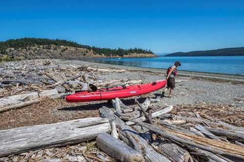 A person pulls a red kayak onto a beach scattered with driftwood, with calm water and hills in the background.