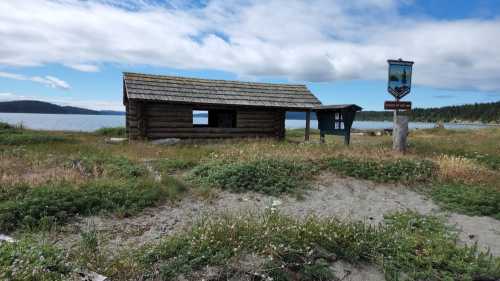 A rustic log cabin near a beach, surrounded by grass and wildflowers, with a signpost nearby and a cloudy sky above.