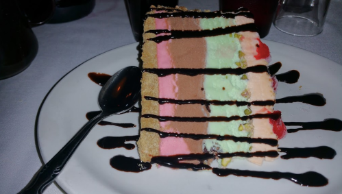 A slice of layered ice cream cake with chocolate, mint, and strawberry flavors, drizzled with chocolate sauce.