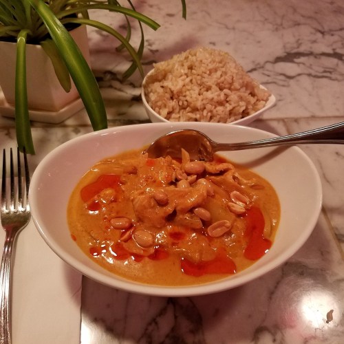 A bowl of chicken curry topped with peanuts and drizzled with sauce, served with a side of brown rice.