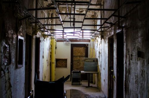 An abandoned hallway with peeling walls, broken ceiling, and a wheelchair in the foreground. Light filters through a window.