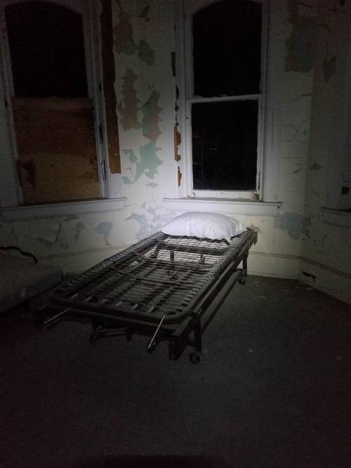A dimly lit room with a bare metal bed frame and a pillow, surrounded by peeling wallpaper and dark windows.