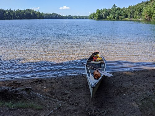 A canoe on a sandy shore by a calm lake, surrounded by lush green trees and a clear blue sky.