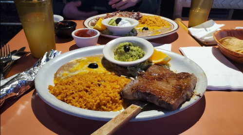 A plate of steak with rice, guacamole, and sides, accompanied by drinks and condiments on a restaurant table.