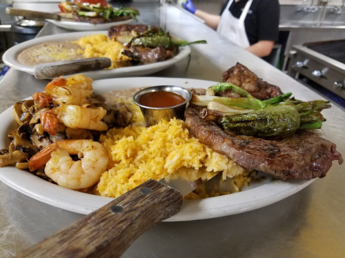 A plate of grilled shrimp, steak, rice, and vegetables, served with a small cup of sauce.