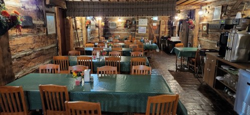Interior of a rustic restaurant with wooden walls, green tablecloths, and rows of wooden chairs set for dining.