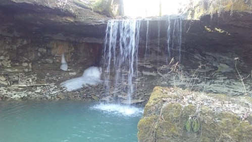 A serene waterfall cascades into a clear pool, surrounded by rocky terrain and mossy edges. Sunlight filters through.