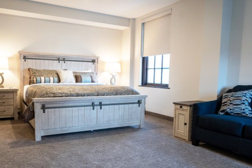 A cozy bedroom featuring a large bed, nightstands, and a blue sofa, with natural light from a window.