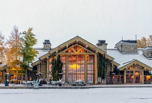 A cozy lodge with large windows, surrounded by snow and autumn trees, creating a serene winter scene.