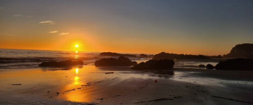 Sunset over a tranquil beach, with golden light reflecting on the water and rocky formations along the shore.