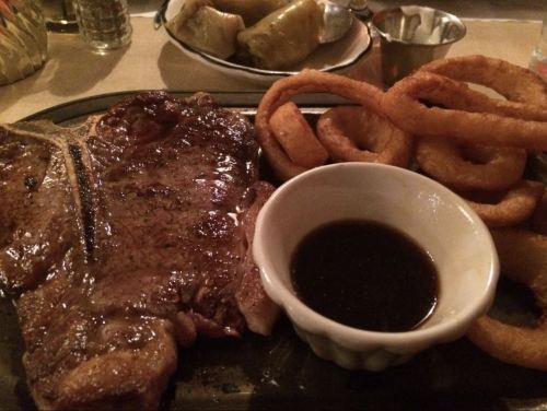 A sizzling steak on a plate with onion rings and a small bowl of sauce.