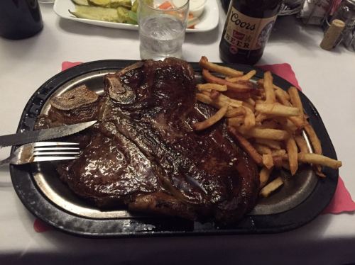 A large steak on a plate with golden fries, accompanied by a glass of water and a beer in the background.