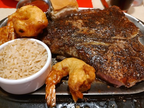 A plate featuring grilled steak, shrimp, rice, a bun, and a side of sauce.