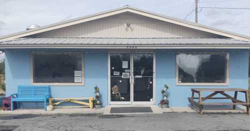 A blue building with a gray roof, featuring a welcoming entrance, outdoor seating, and flower pots by the windows.