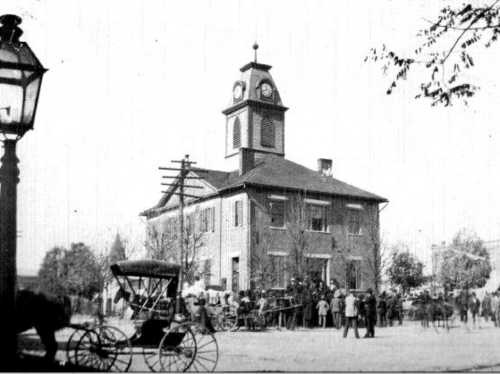 Historic black-and-white photo of a clock tower building with a crowd gathered outside, horse-drawn carriage in foreground.