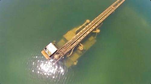 Aerial view of a long structure extending into clear water, possibly a pier or oil rig, with sunlight reflecting on the surface.