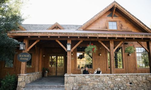 A rustic wooden restaurant exterior with a stone foundation, featuring hanging plants and two people seated outside.