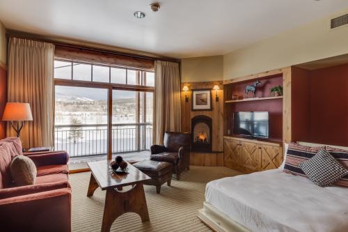 Cozy hotel room with a fireplace, large window, and mountain view, featuring a bed, sofa, and wooden furniture.