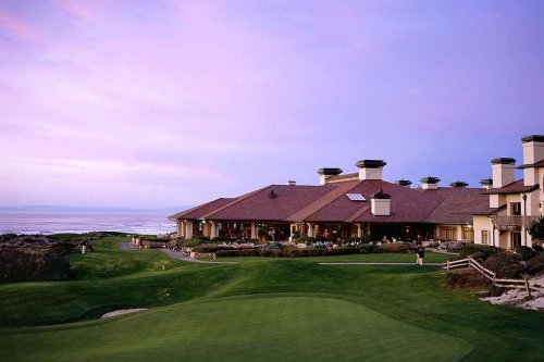 A scenic view of a golf club building at sunset, overlooking a lush green lawn and the ocean in the background.