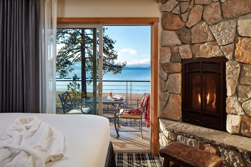 Cozy room with a view of a lake, featuring a stone fireplace and a balcony with chairs overlooking the water.