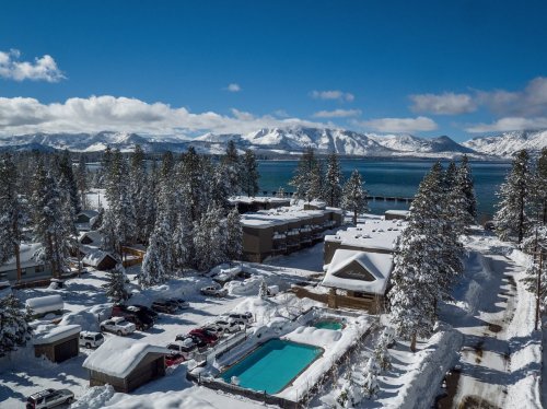 Aerial view of a snowy landscape with a lake, mountains, and a resort featuring a pool and parked cars.