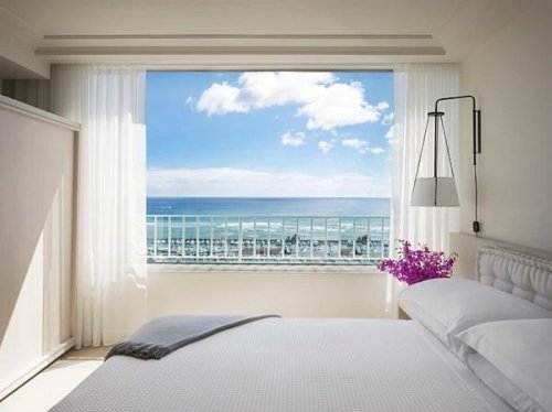 Bright bedroom with a white bed, sheer curtains, and a stunning ocean view through a large window.
