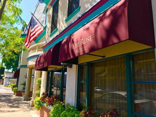 Exterior of The Rose Hotel with an awning, American flag, and greenery along the sidewalk on a sunny day.
