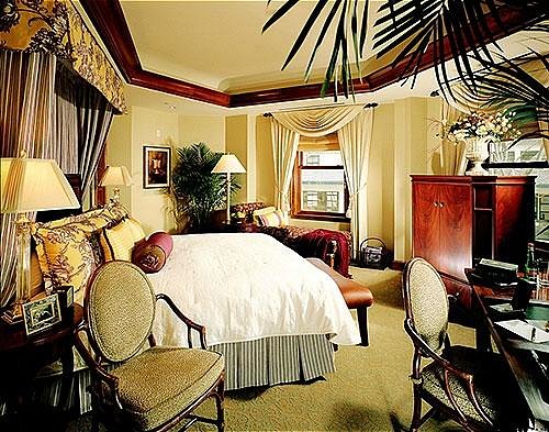 Luxurious hotel room with a king bed, elegant decor, two chairs, and a large window with drapes.