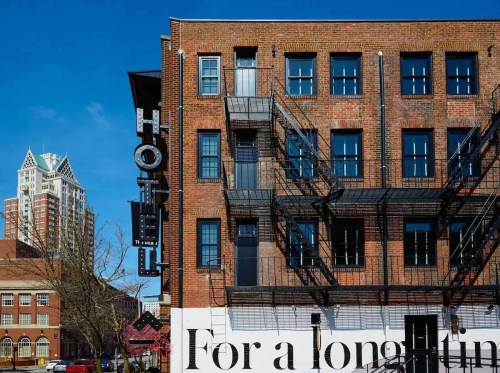 A brick building with fire escapes and a hotel sign, featuring a mural that reads "For a long time." Clear blue sky.