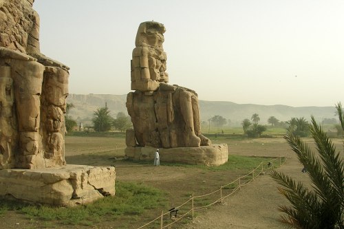 Two massive ancient statues sit in a desert landscape, with palm trees and mountains in the background.