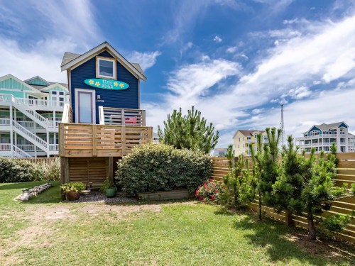 A blue beach house with a deck surrounded by greenery and colorful flowers under a bright, cloudy sky.