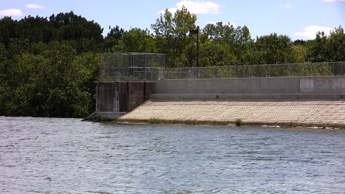 A water dam with a sloped concrete surface, surrounded by trees and a blue sky with scattered clouds.