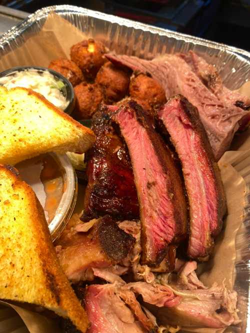 A tray of barbecue featuring sliced meats, cornbread, coleslaw, and fried cornbread balls.