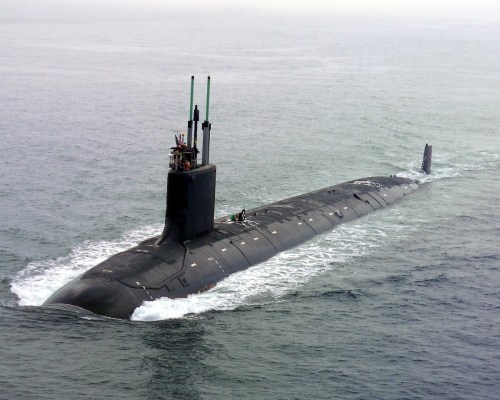 A submarine navigates through calm waters, with its conning tower and antennas visible above the surface.
