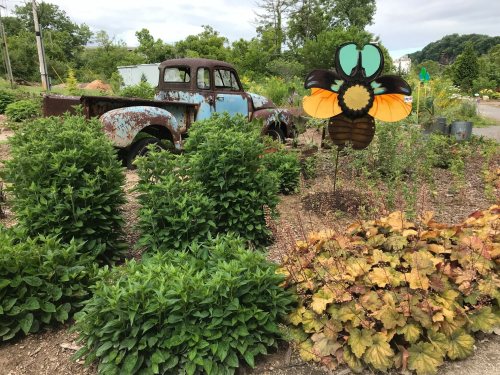An old, rusty truck is surrounded by lush greenery and a colorful bee sculpture in a garden setting.