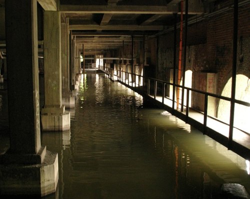 Dimly lit underground space with water reflecting light, supported by concrete pillars and graffiti on the walls.