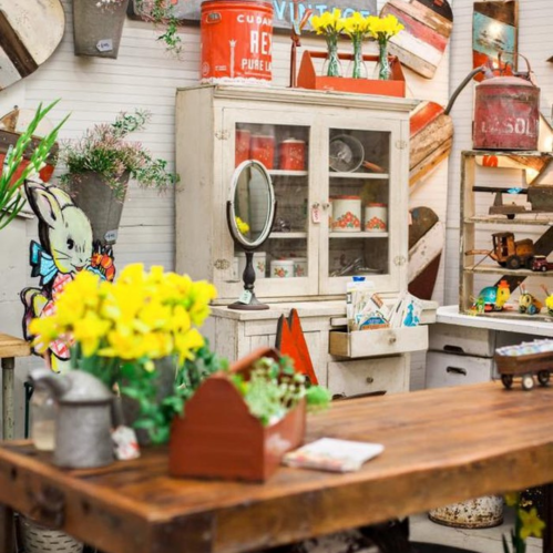 A vintage-style room with a wooden table, flowers, a cabinet, and various decorative items on the walls.