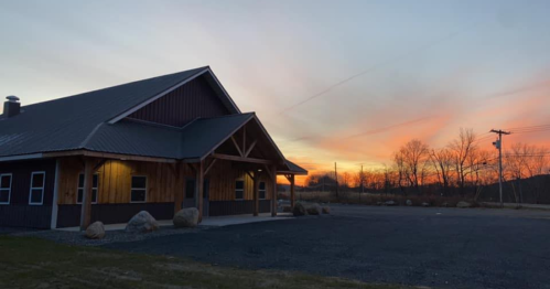 A rustic building at sunset, with a colorful sky and trees in the background, casting a warm glow on the structure.