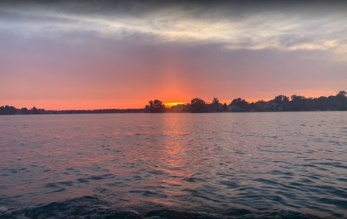 A serene lake at sunset, with vibrant orange and pink hues reflecting on the water and silhouettes of trees and houses in the distance.