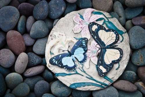 A decorative stone featuring painted butterflies and flowers, resting on a bed of smooth, colorful pebbles.