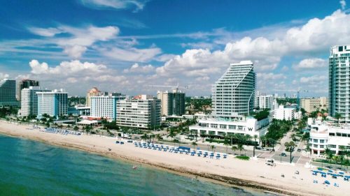 Aerial view of a beachside city with modern buildings, sandy shore, and blue umbrellas under a partly cloudy sky.