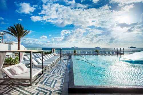 A luxurious poolside view with lounge chairs, palm trees, and a bright blue sky over the ocean.