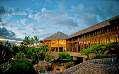 A tropical resort with a thatched roof, surrounded by palm trees and a serene water feature under a colorful sky.