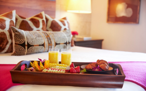 A wooden tray with two glasses of juice, croissants, and fresh fruit on a bed with decorative pillows.