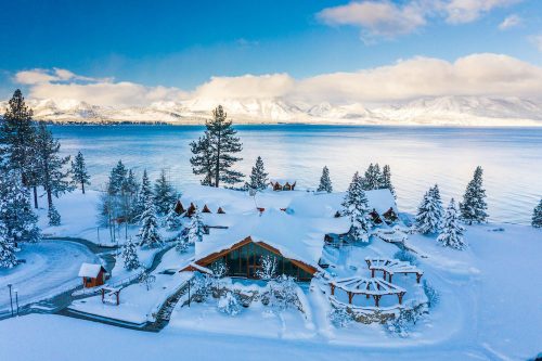 A snowy landscape featuring a cozy lodge by a lake, surrounded by evergreen trees and mountains under a clear blue sky.